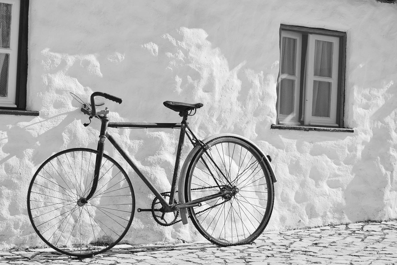 A dark bicycle rests against a pale textured wall between two windows on a cobbled surface.