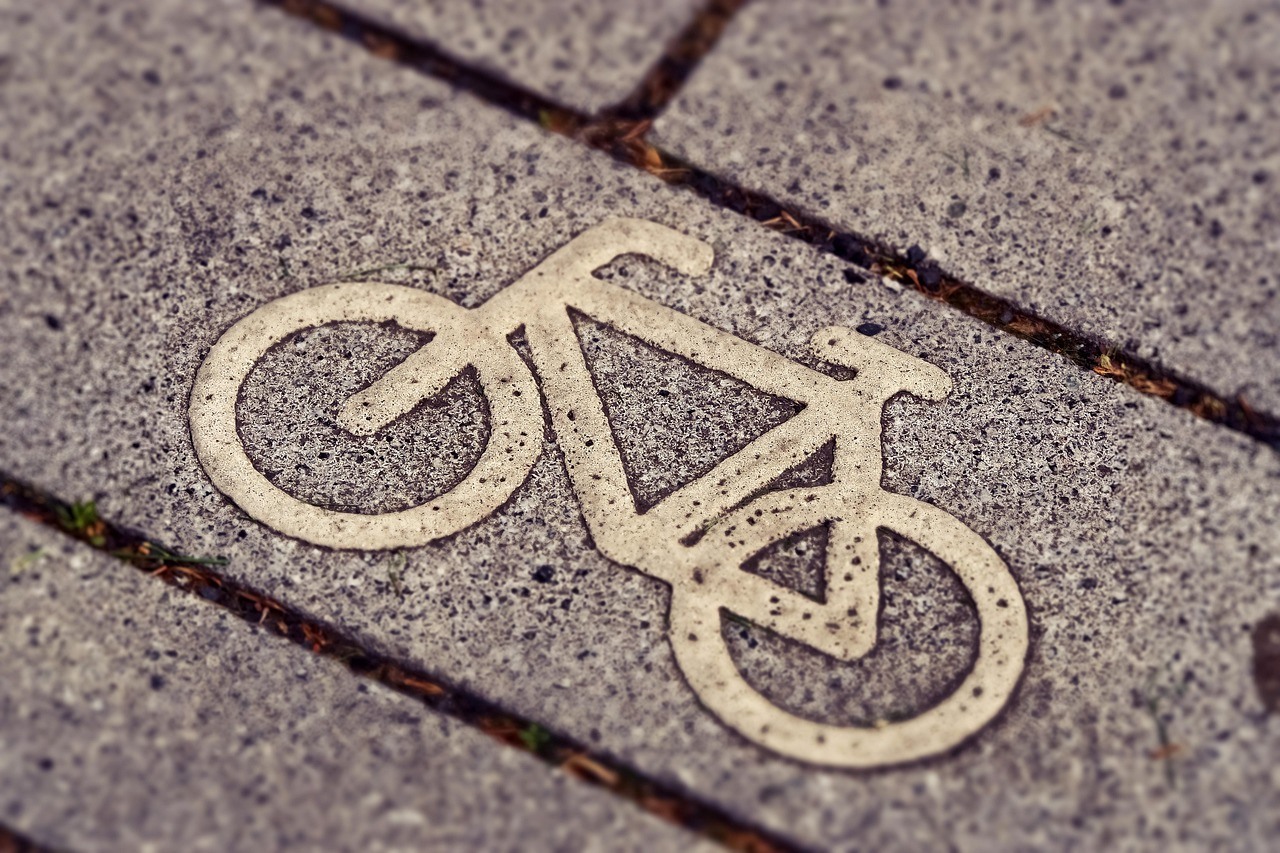 A pale bicycle symbol is embedded in coarse pavement beside dark joints and scattered moss.