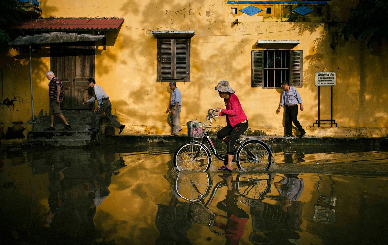 A cyclist in a bright red top crosses a large puddle beside a yellow building as pedestrians move nearby.