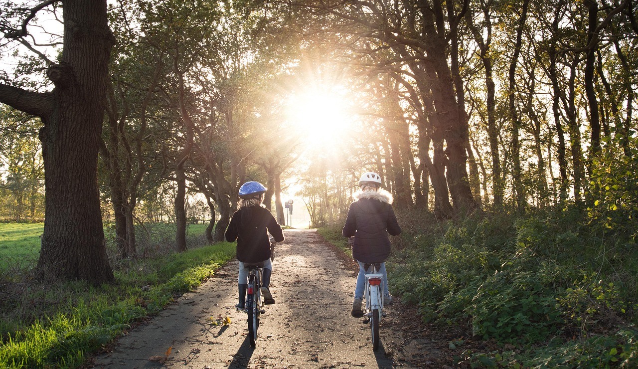 Two cyclists ride away along a wooded dirt path toward low sunlight shining through the trees.